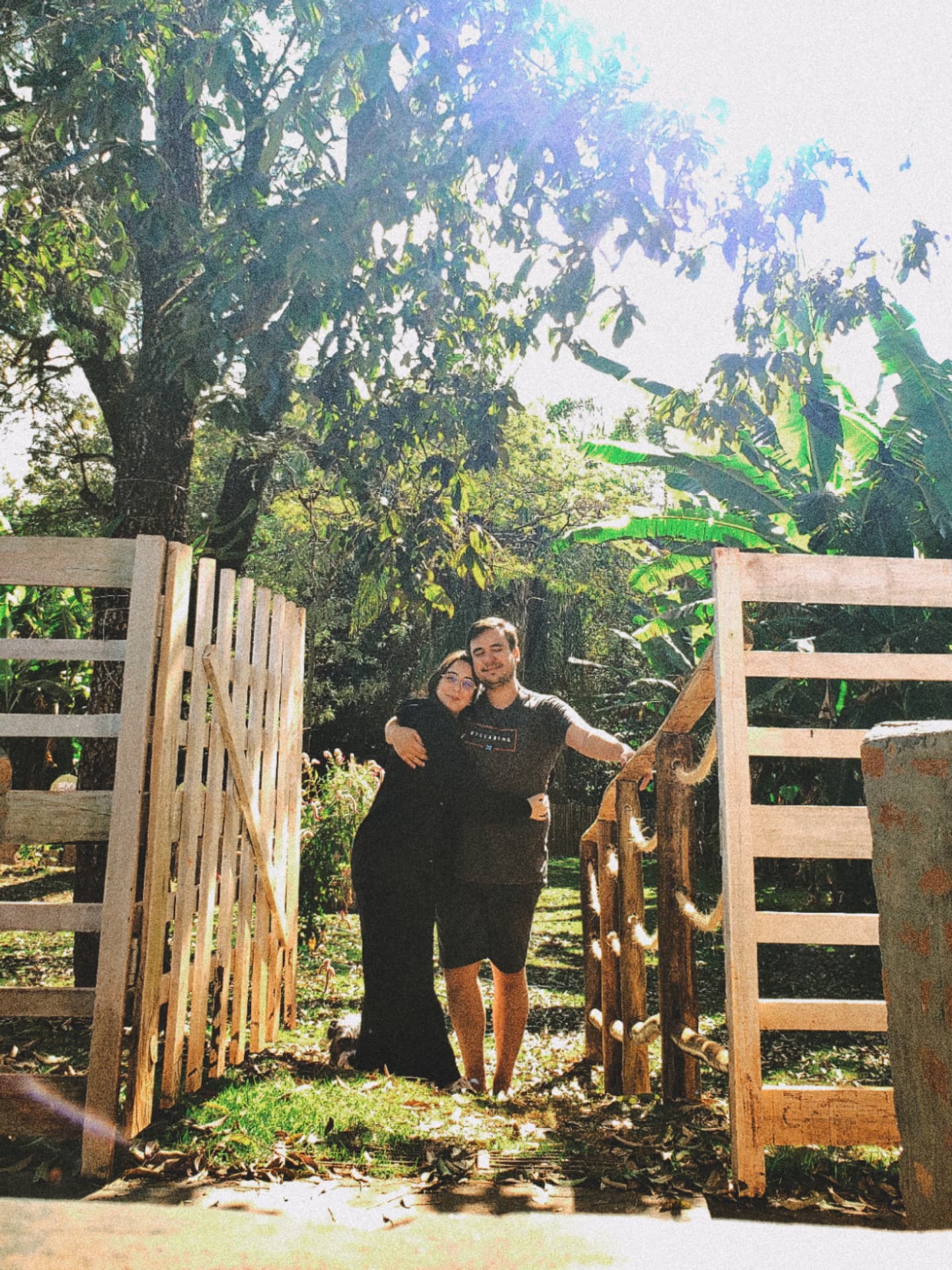 Bárbara e William abraçados no portão de madeira em meio ao verde