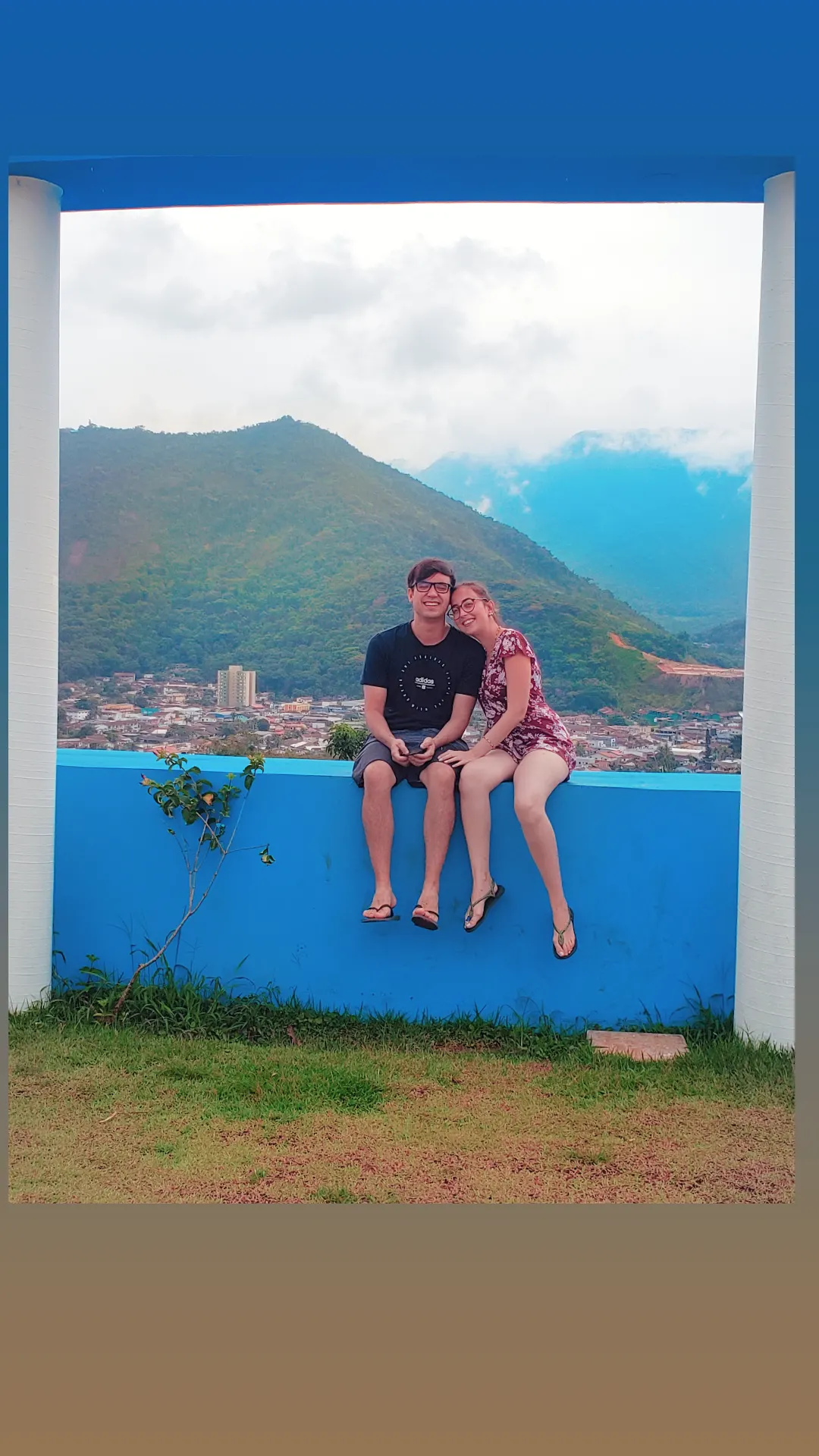 Bárbara e William sentados no muro azul com a serra ao fundo